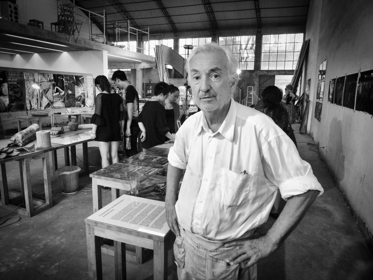 A gray-haired man in a white shirt and workshop apron stands with his hands on his hips inside an architectural studio