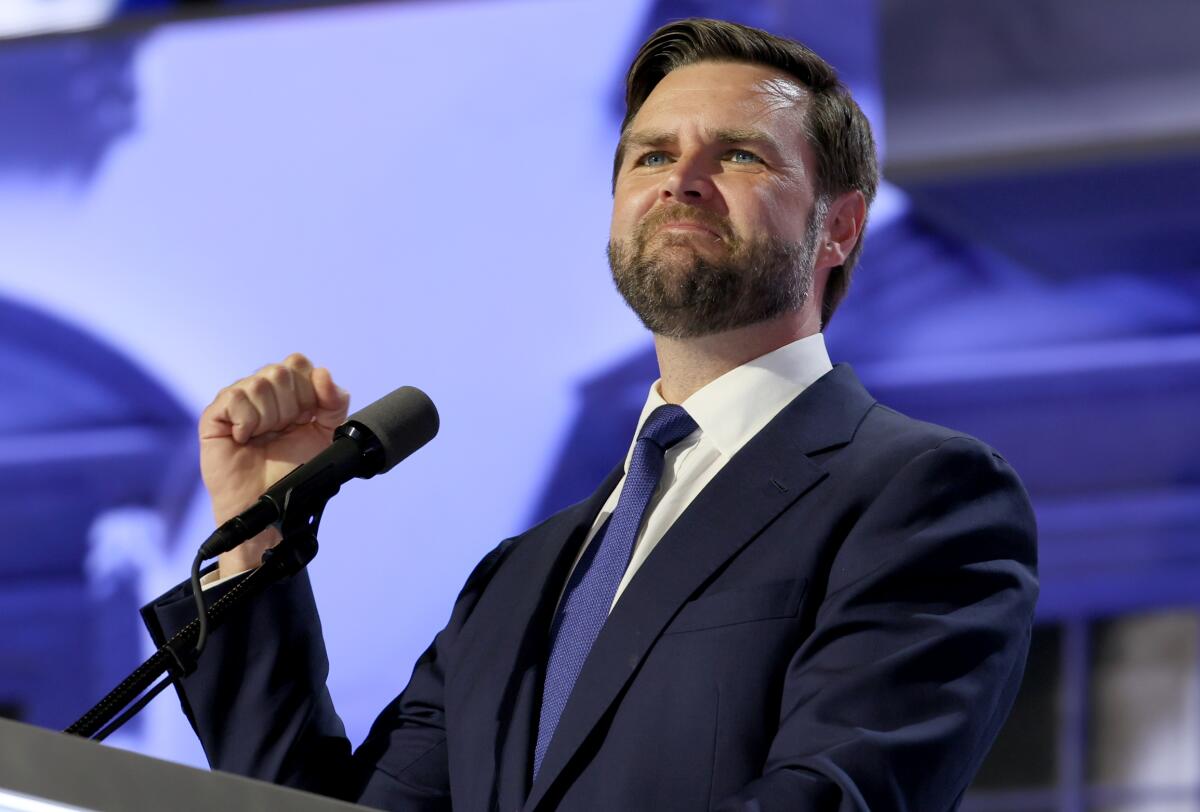 A man with dark hair and beard, in dark suit and blue tie, holds a fisted hand while standing before a microphone 