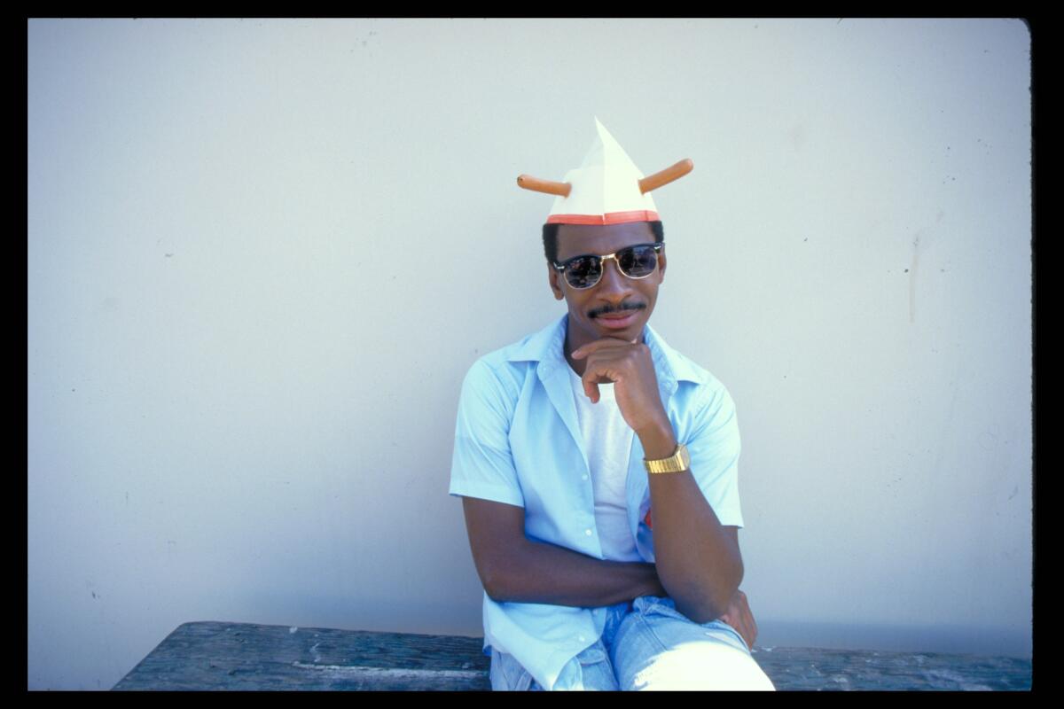 Director Robert Townsend in a blue shirt and hat with his chin on his hand.