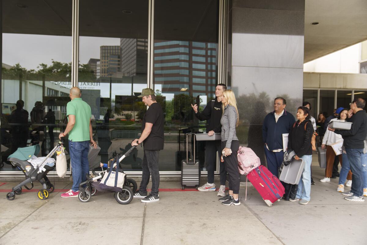 People standing in line, many with strollers or luggage, on the sidewalk outside a building with large windows