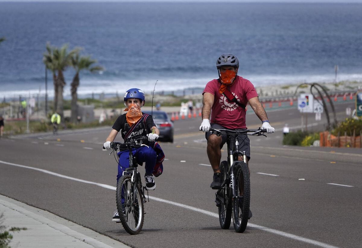 Wearing cloth face coverings and gloves, Neal Tricarico and son Anthony ride along Coast Highway in Encinitas on April 5. All beaches in San Diego County have been closed because of COVID-19, but could begin to reopen as of Monday. Face coverings will be required countywide starting May 1.