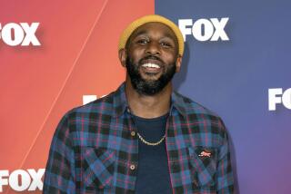 A man in a yellow beanie, blue flannel shirt and gold chain tilting his head up and smiling