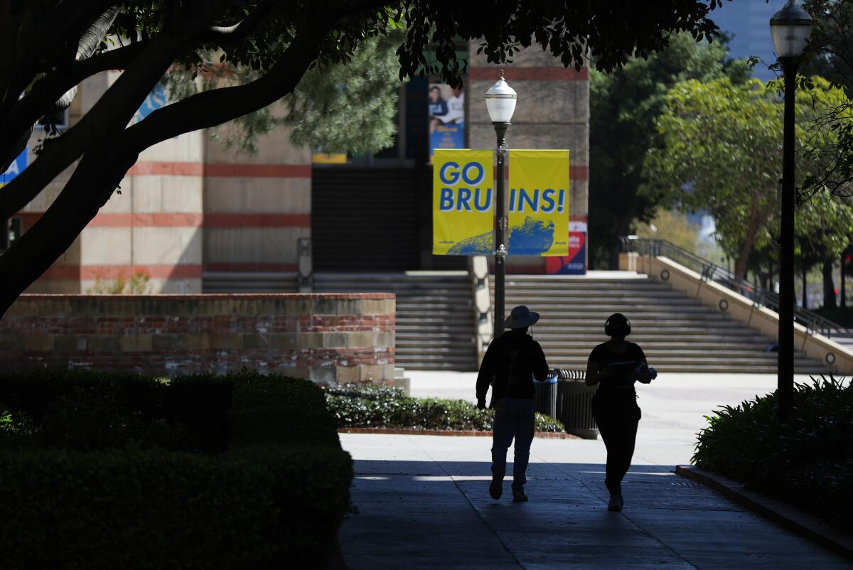 Silhouette of two people walking beneath a tree and in front of a "Go, Bruins" sign on a campus.
