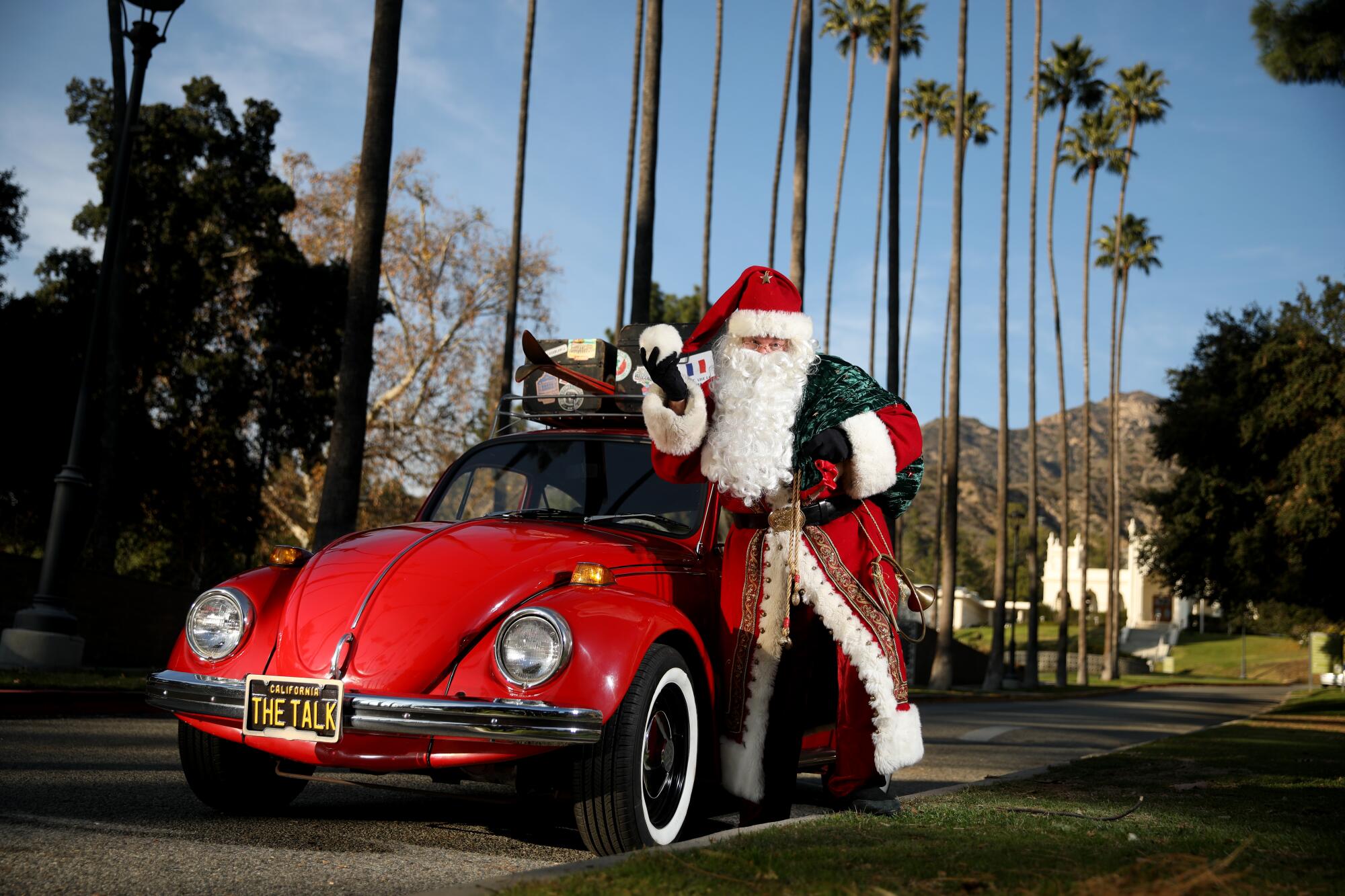 A man dressed as Santa Claus leans against a red Volkswagen Beetle car.