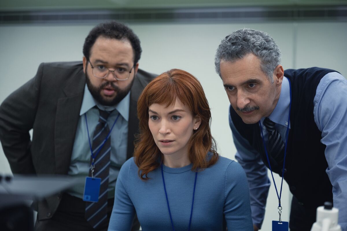 A woman sits at a computer with two men looking over her shoulders at the screen in a scene from "Severance."