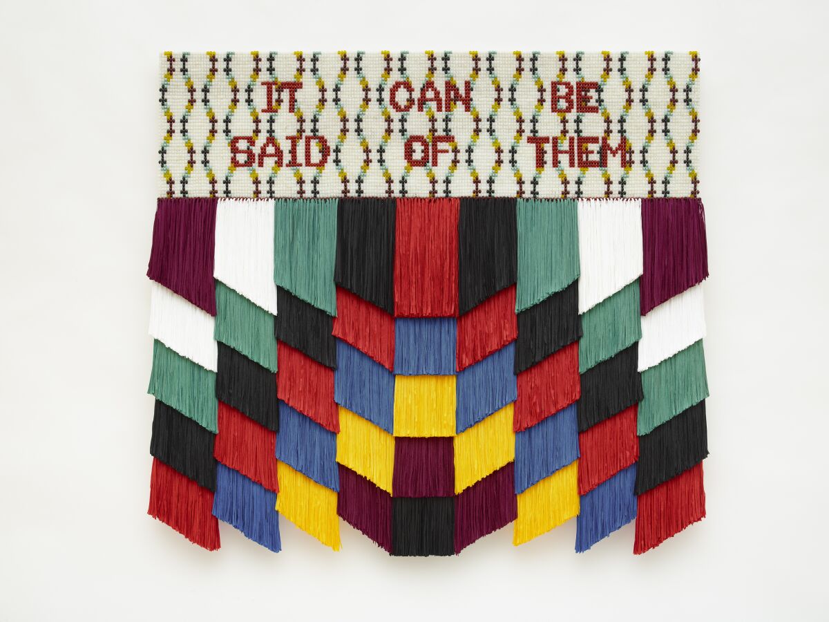 A multimedia art work features fringe in a geometric pattern topped by the phrase "It Can Be Said of Them" in beads.
