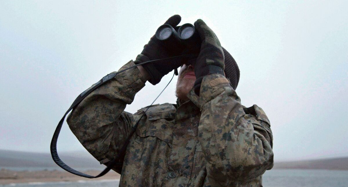 A scientist dressed in warm camouflage gear looks through binoculars.
