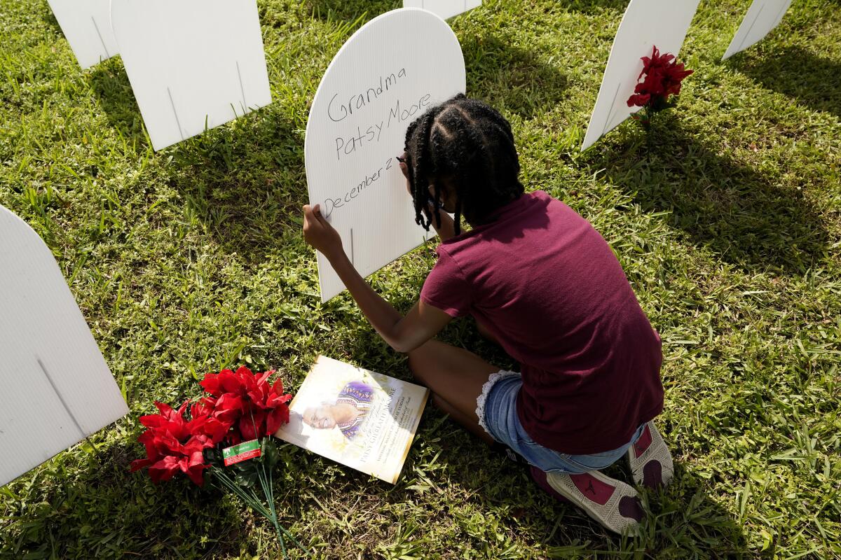 A girl kneels on grass as she writes the name of her grandmother on a cardboard headstone.