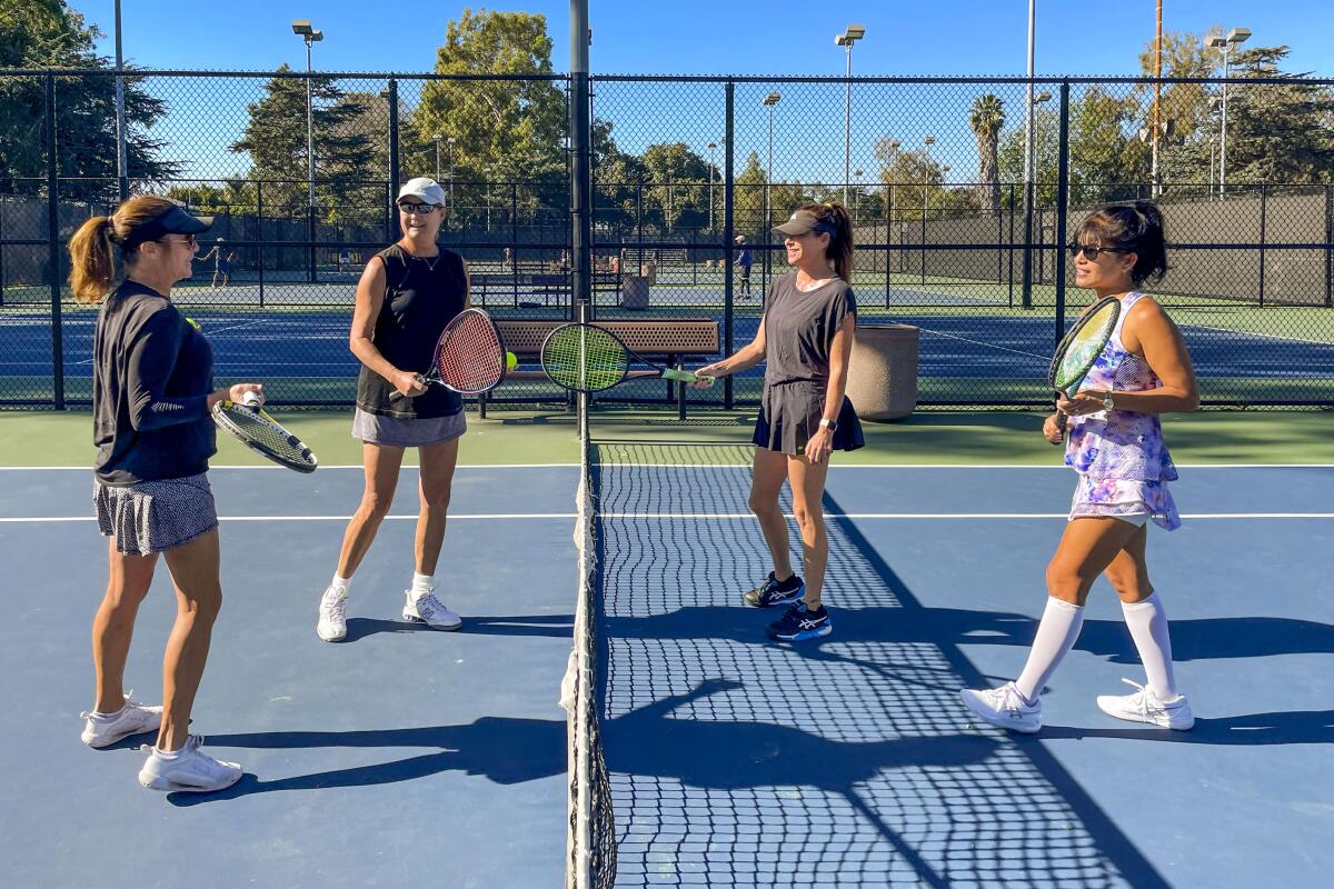 Four women stand near on either side of the net of a tennis court. 