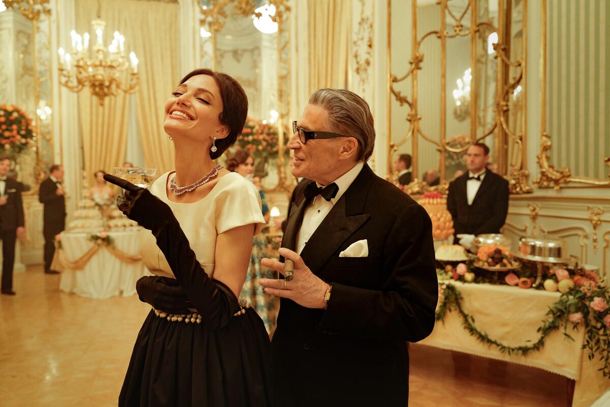 A woman carrying champagne walks next to a man in a tuxedo holding a cigar in an ornate room