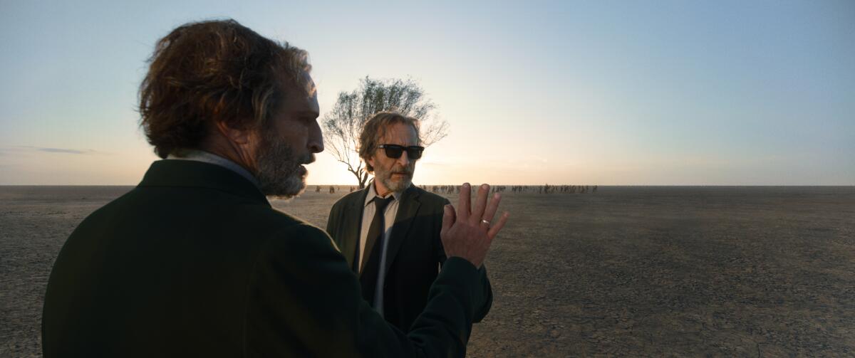 A man stands in a desert-like landscape facing his mirror image.