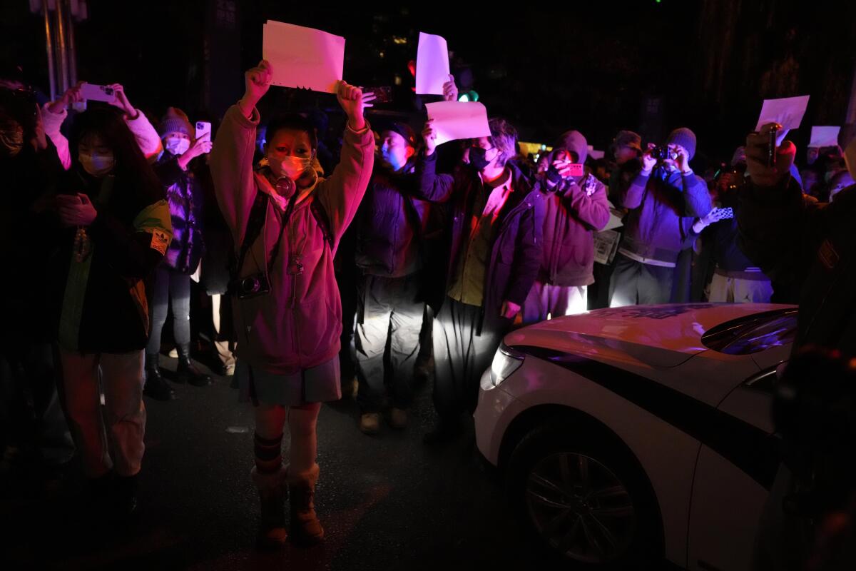 A crowd of protesters in masks and winter jackets hold up blank papers in a night scene illuminated by a car's headlights.