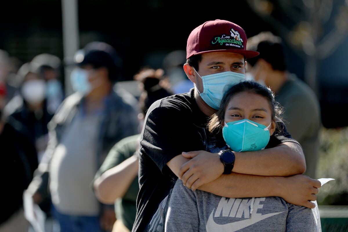 A man playfully hugs his sister as they wait in a line. Both wear masks.
