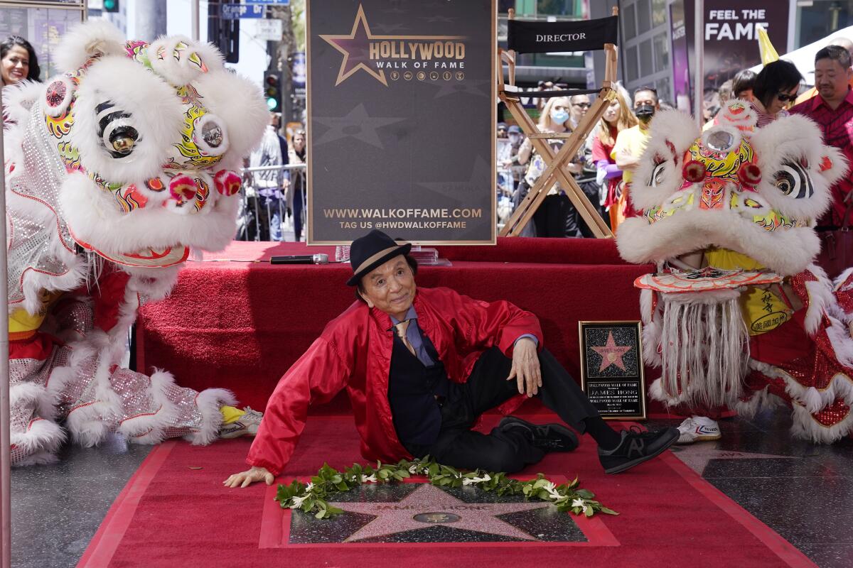 An actor sits on red carpet next to his star on Hollywood Boulevard, with Chinese lion dancers behind him.