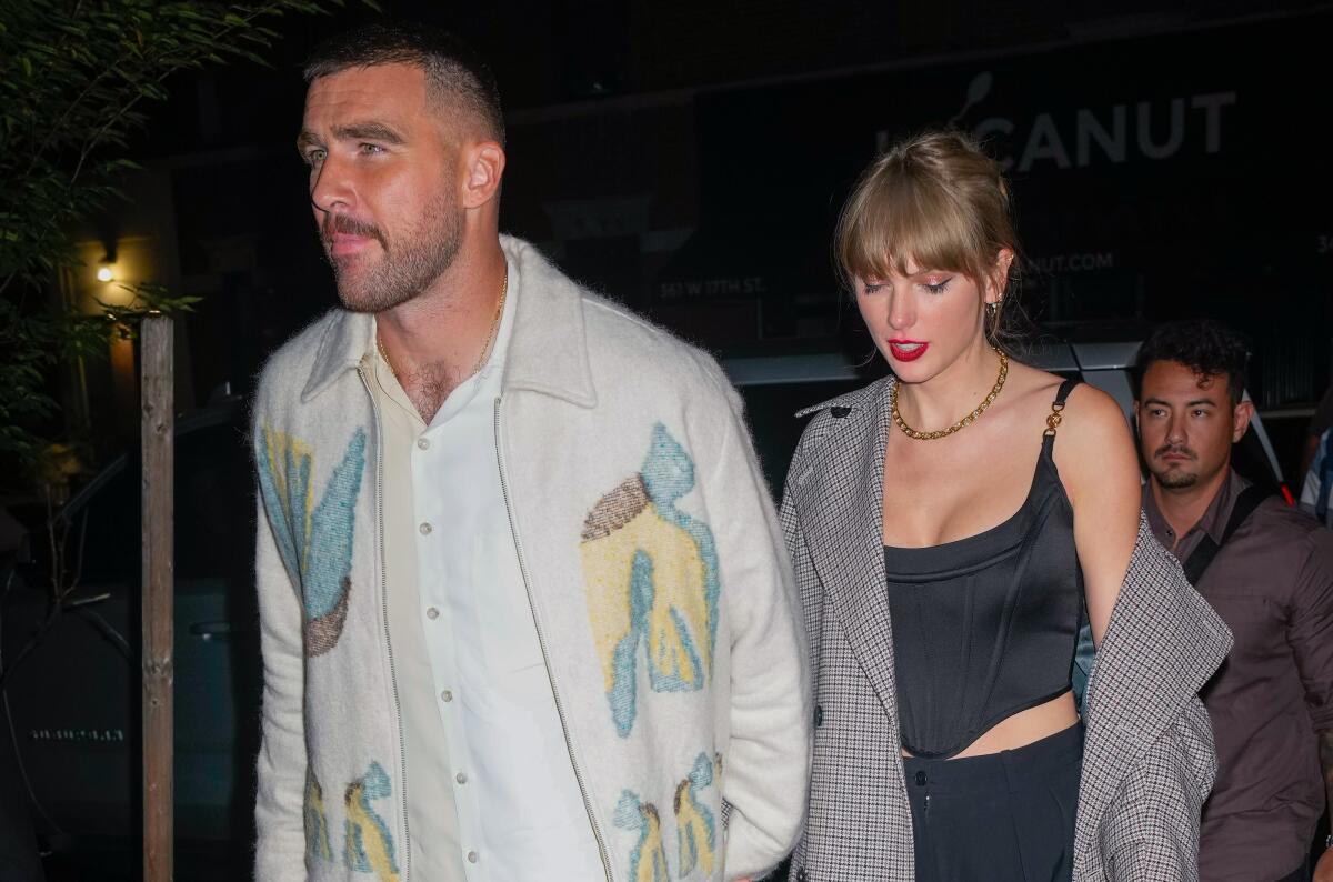 Travis Kelce, in a cardigan over a white shirt, holds hands with Taylor Swift who wears a plaid coat that's off one shoulder