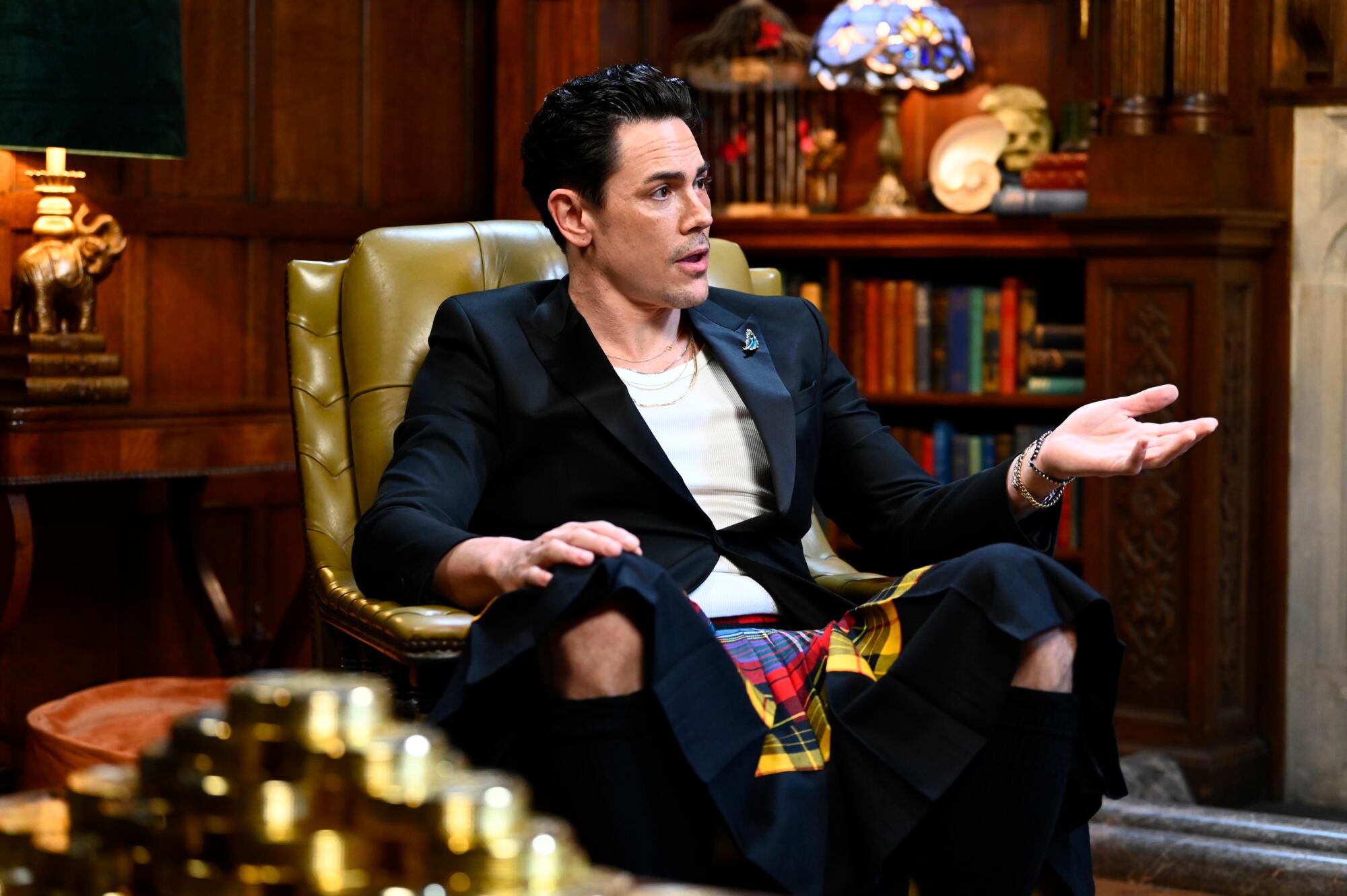 A man in a black tuxedo jacket, white shirt and kilt sits on a green leather chair.