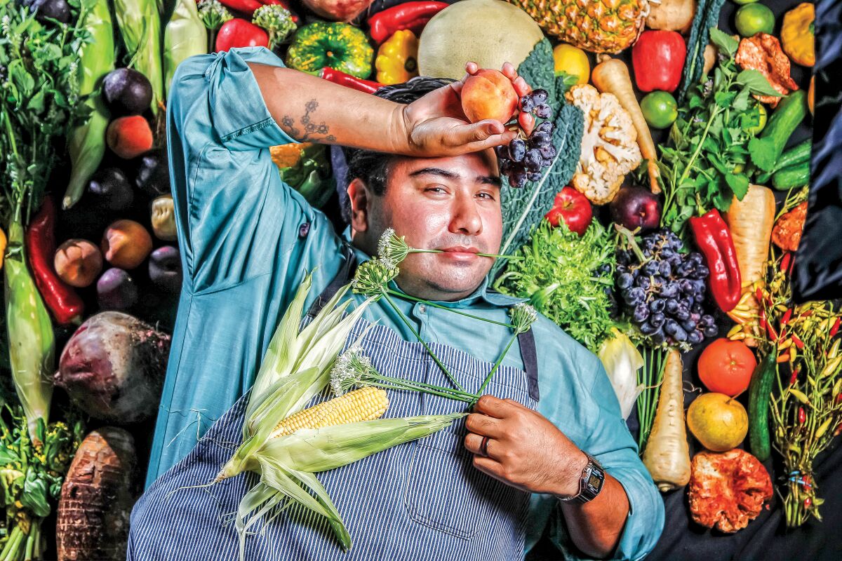 A chef poses playfully on a backdrop of vegetables.