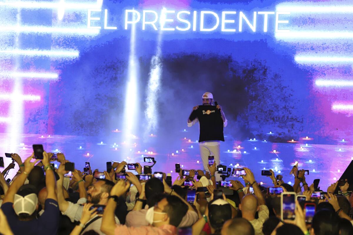 A man in a white cap holds up a black shirt onstage as crowds take cellphone photos of him 