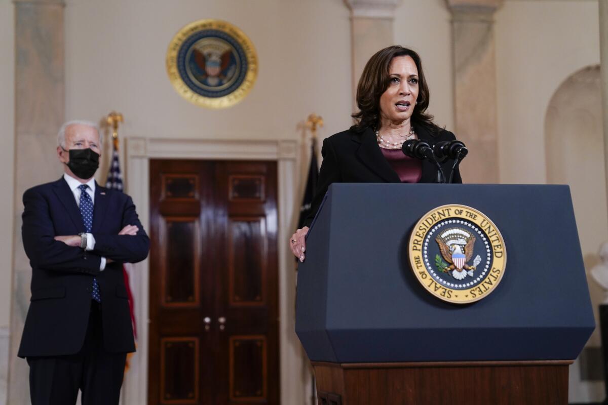 Vice President Kamala Harris speaking at a White House lectern as President Biden stands nearby in a mask