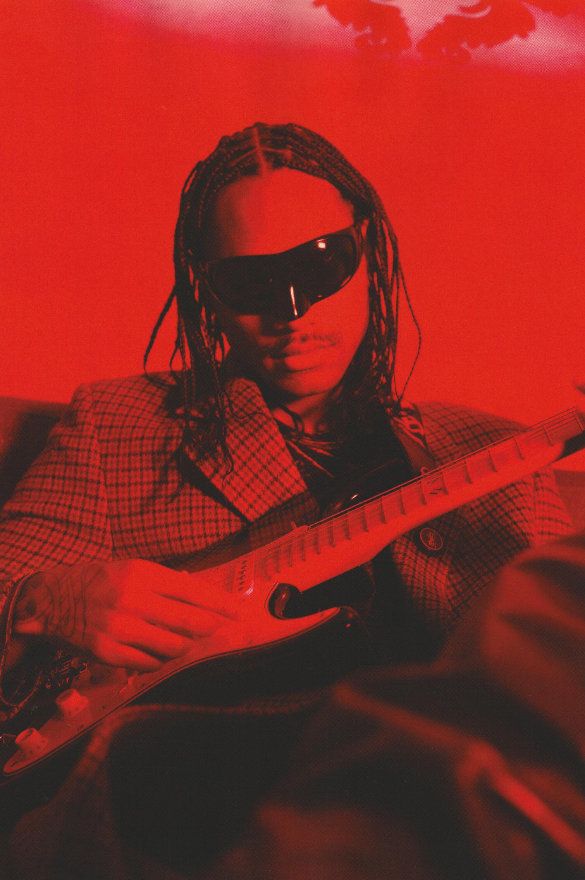 A man in sunglasses, seated, cradles an electric guitar