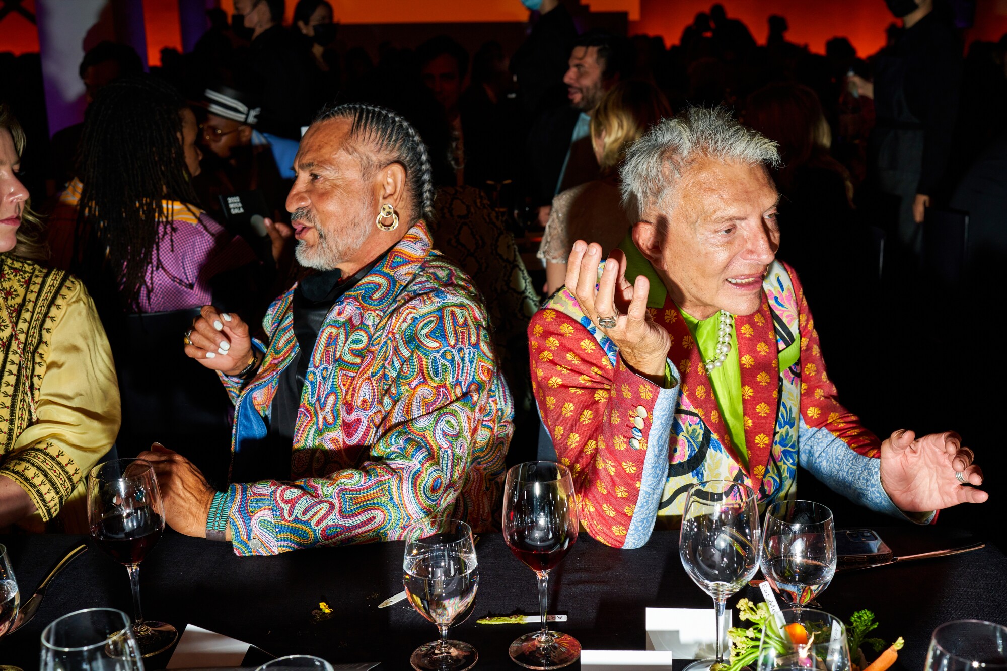 Two men in colorful outfits chat with people at a table.