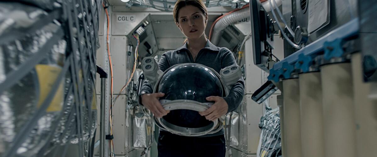 A female astronaut, holding a helmet, looks quizzically into middle distance.