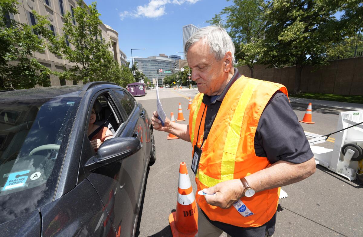 A man in a bright-colored vest accepts a ballot from a voter at a drive-up polling location.