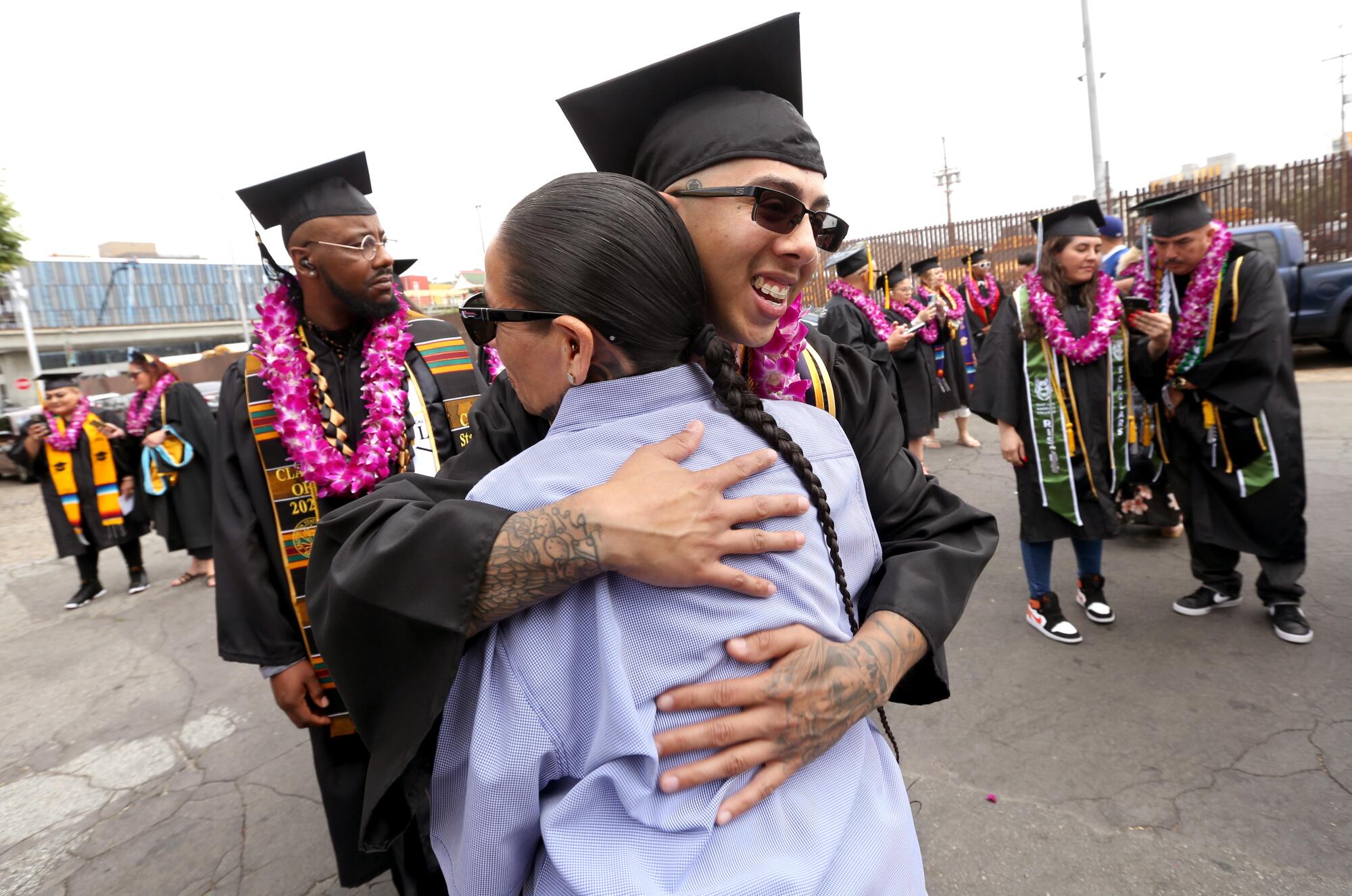 Wearing a graduation gown and cap, Jessi Fernandez faces the camera as he hugs another person