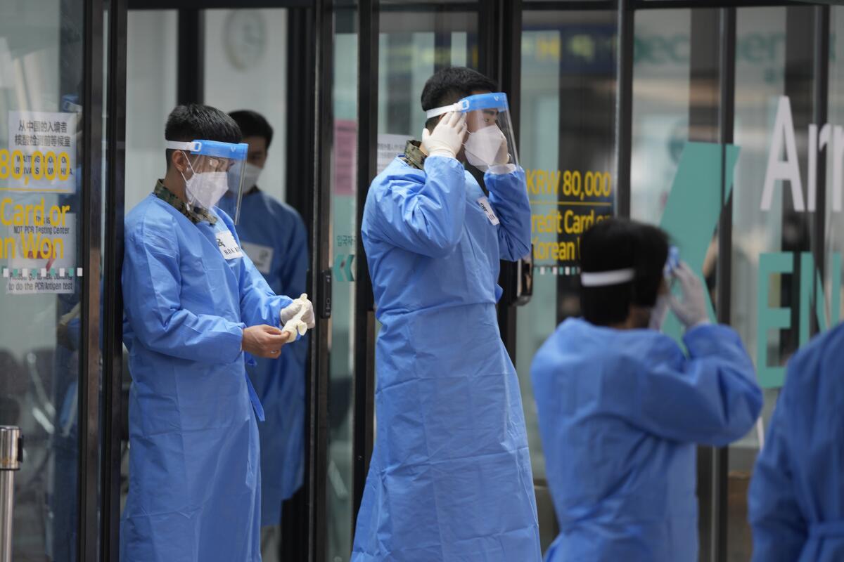 People with face shields and blue protective suits walk outside a building 