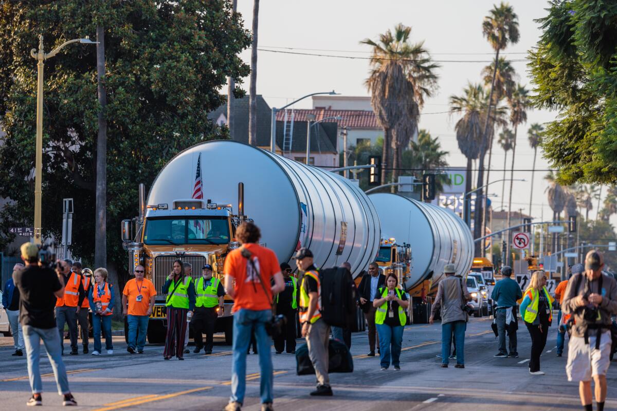 Two large white solid rocket motors are hauled by trucks as people stand nearby.