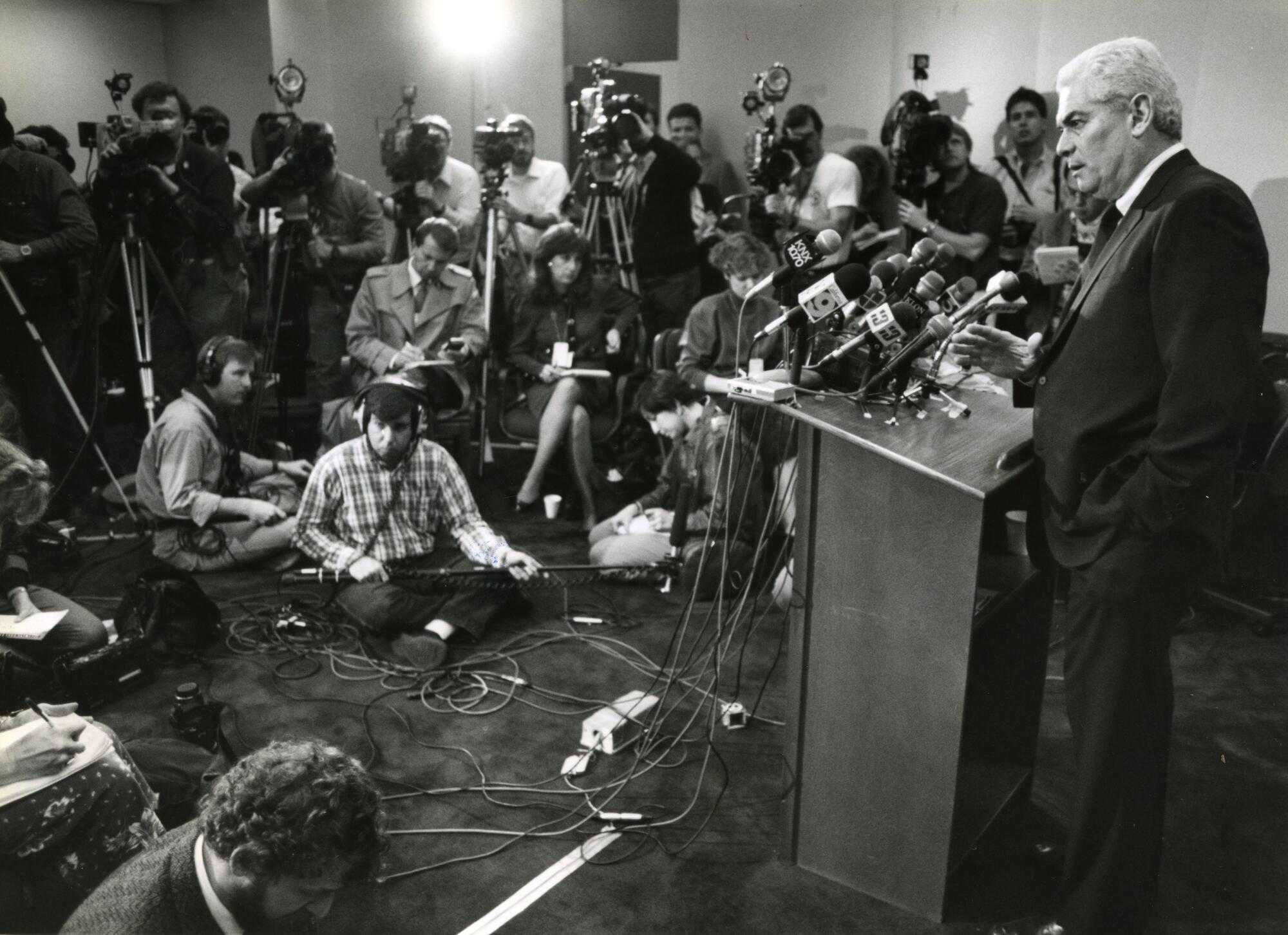 A man addresses a room crowded with journalists, cameras and microphones.