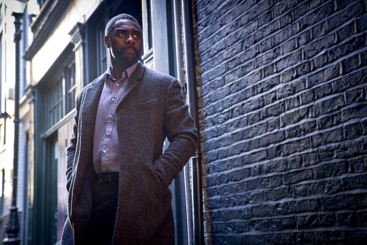 Idris Elba in a suit stands in shadowed light outdoors.