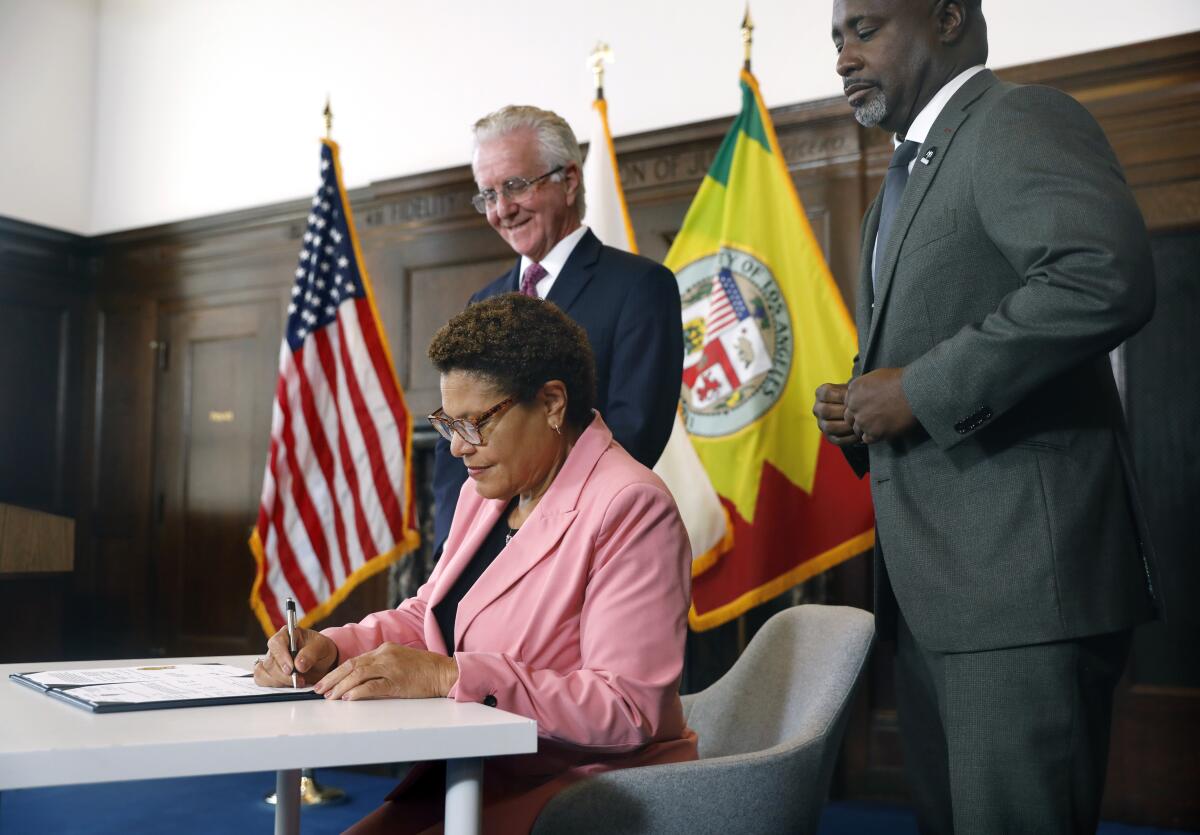 Mayor Karen Bass signs a document seated at a table next to standing colleagues 