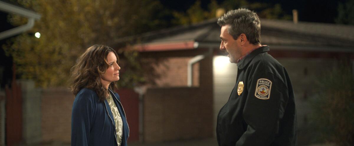 A woman in a robe and a man in a police uniform look at each other outside a home.