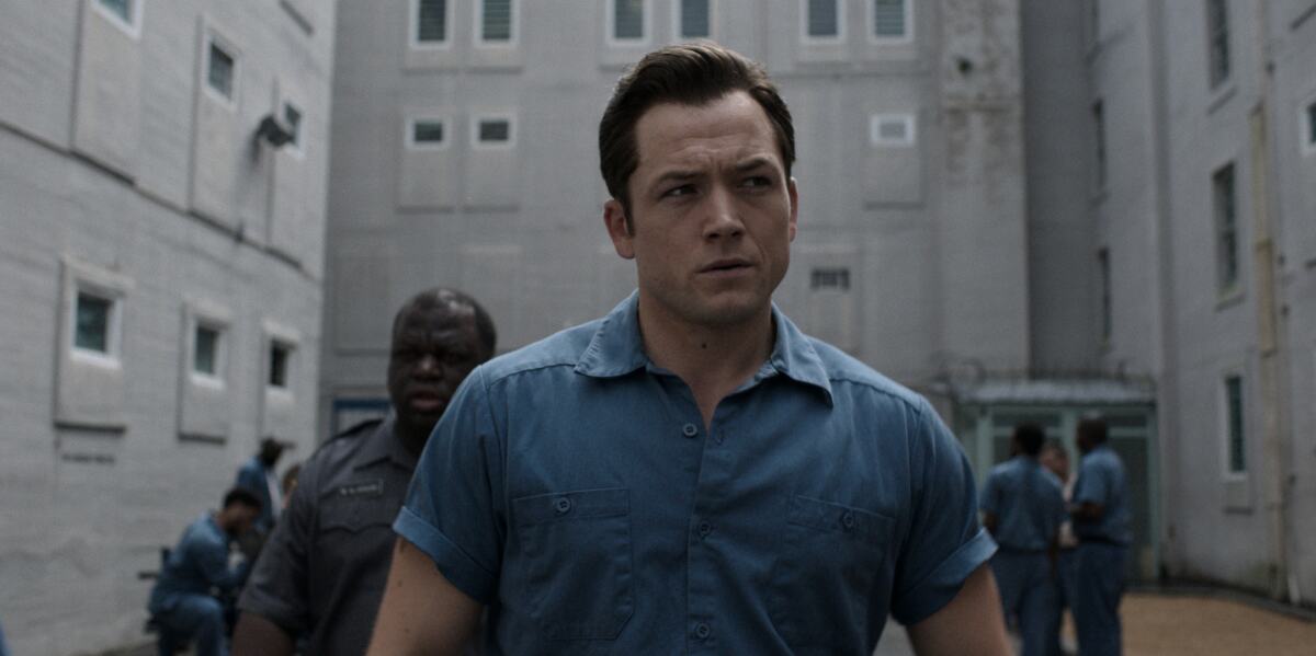 Man looking serious in yard of a prison, with a guard behind him
