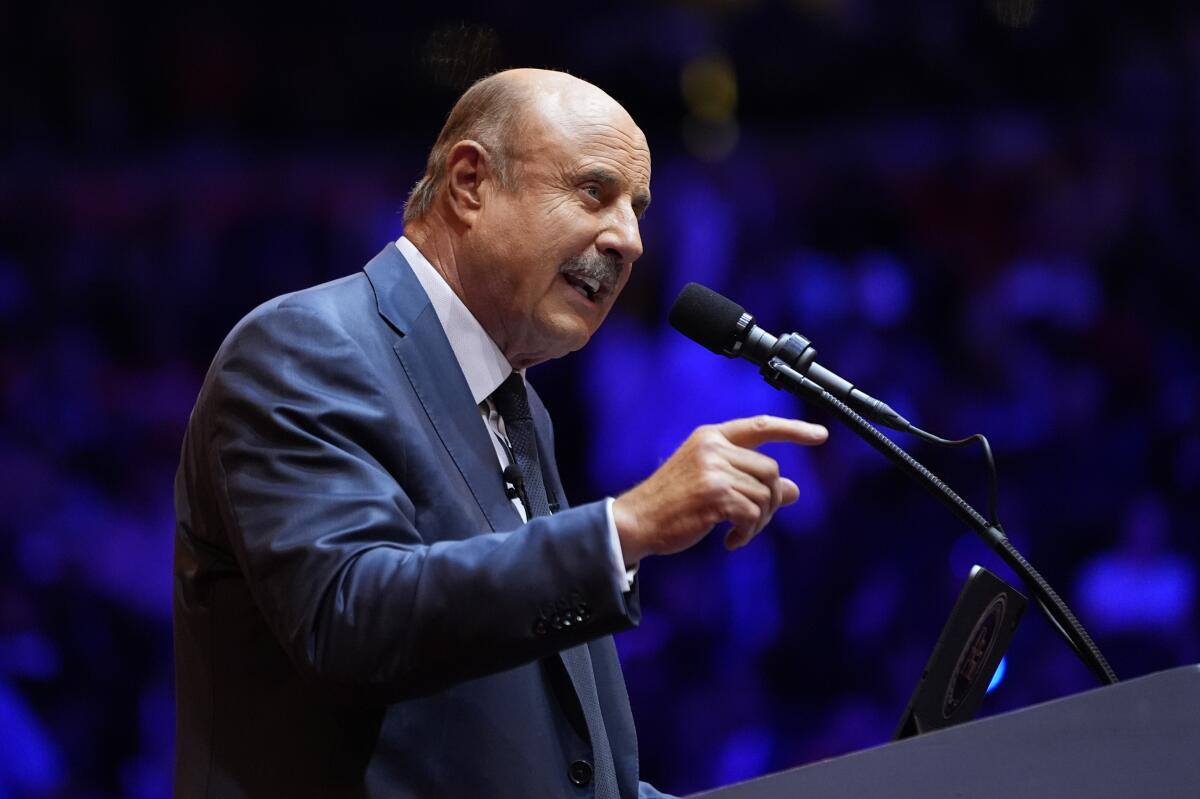 Dr. Phil, in a blue suit and tie, points as he speaks into a microphone at a lectern.
