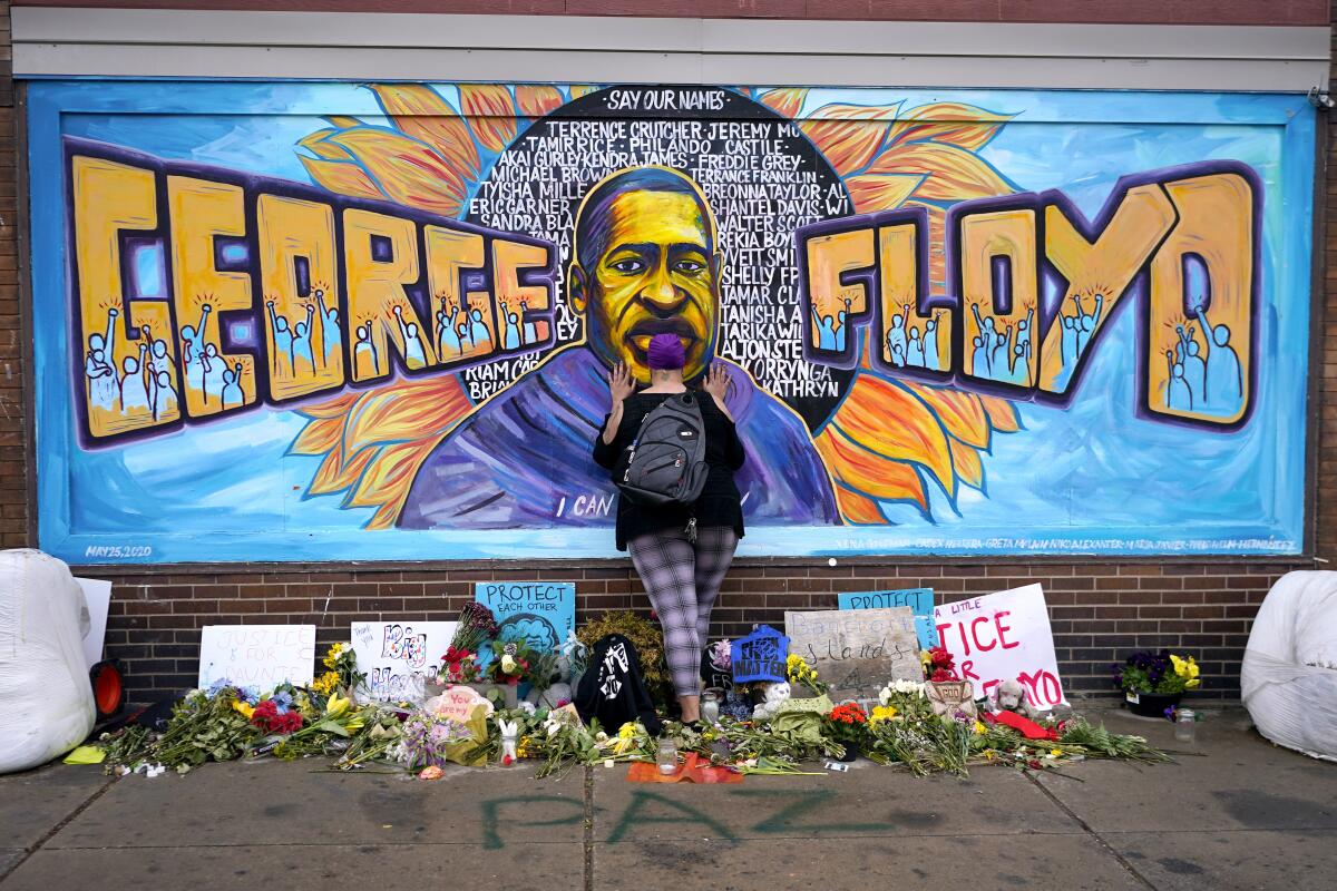 Woman standing in front of memorial for George Floyd set with flowers on the ground and a mural of Floyd with his name 