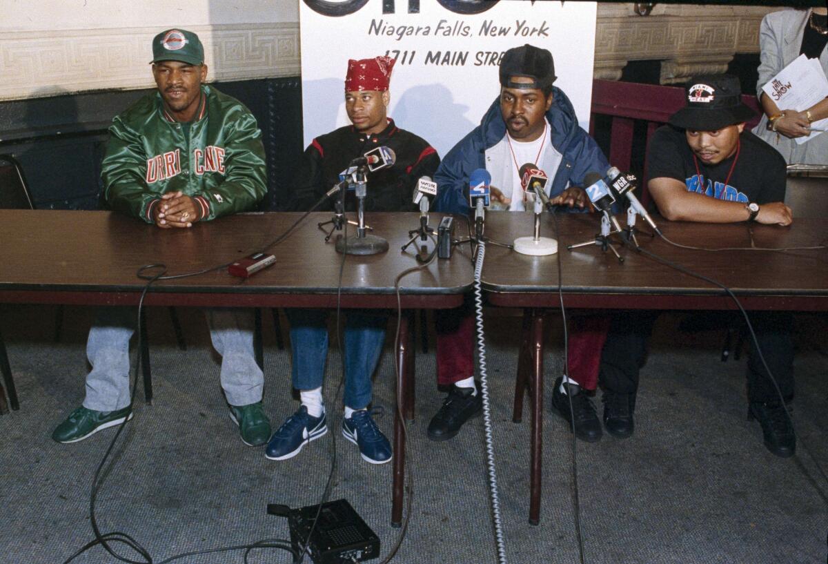 The members of 2 Live Crew sit behind two tables with several microphones