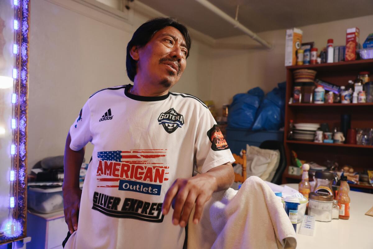A man wearing a T-shirt covered with ads stands next to a kitchen table