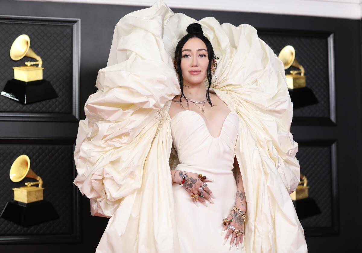Noah Cyrus wears an ivory gown with a cape that bubbles high behind her head in front of a backdrop of Grammys