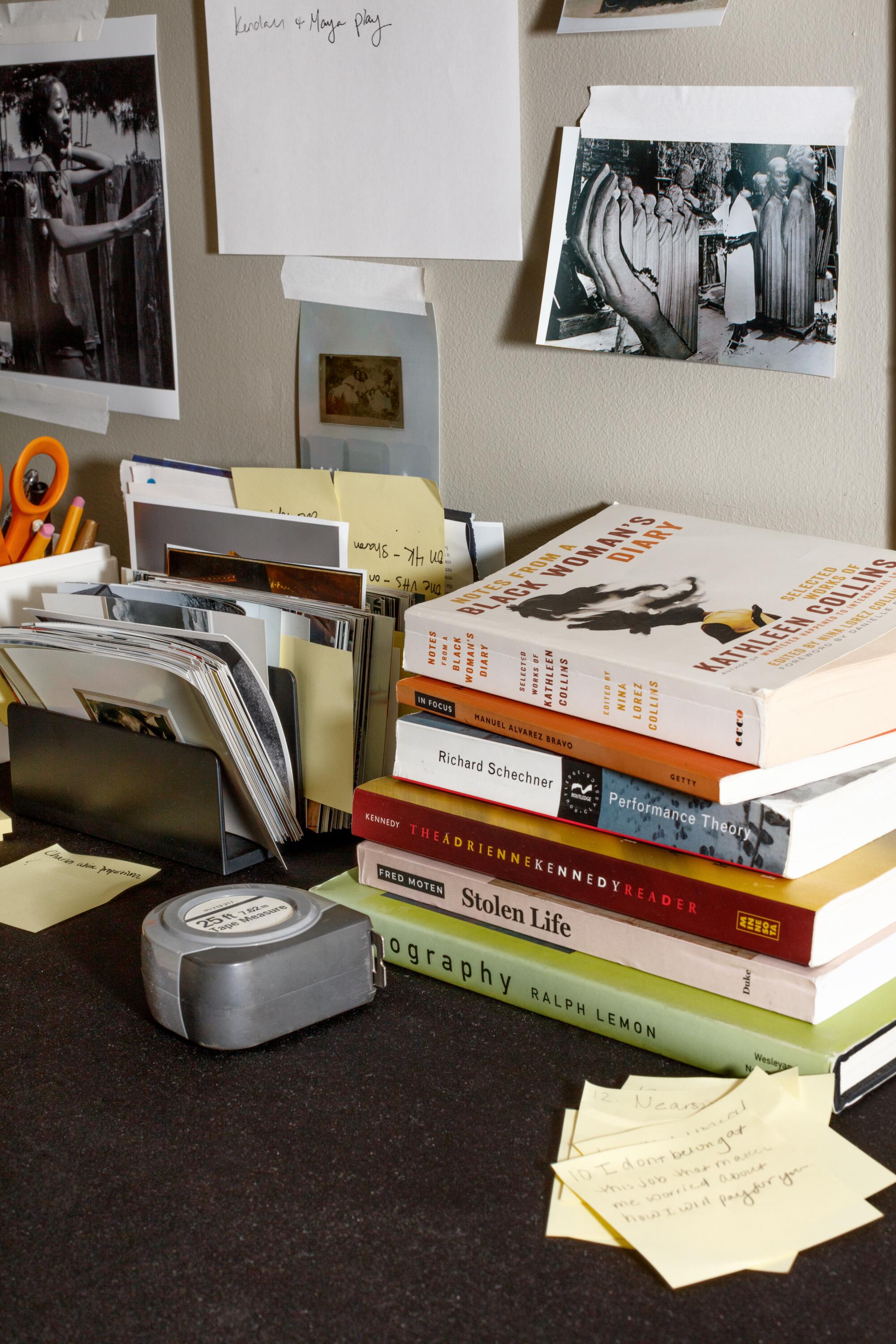 Detail view of a desk with stacked books, measuring tape, yellow post-it notes, and images taped to the wall.