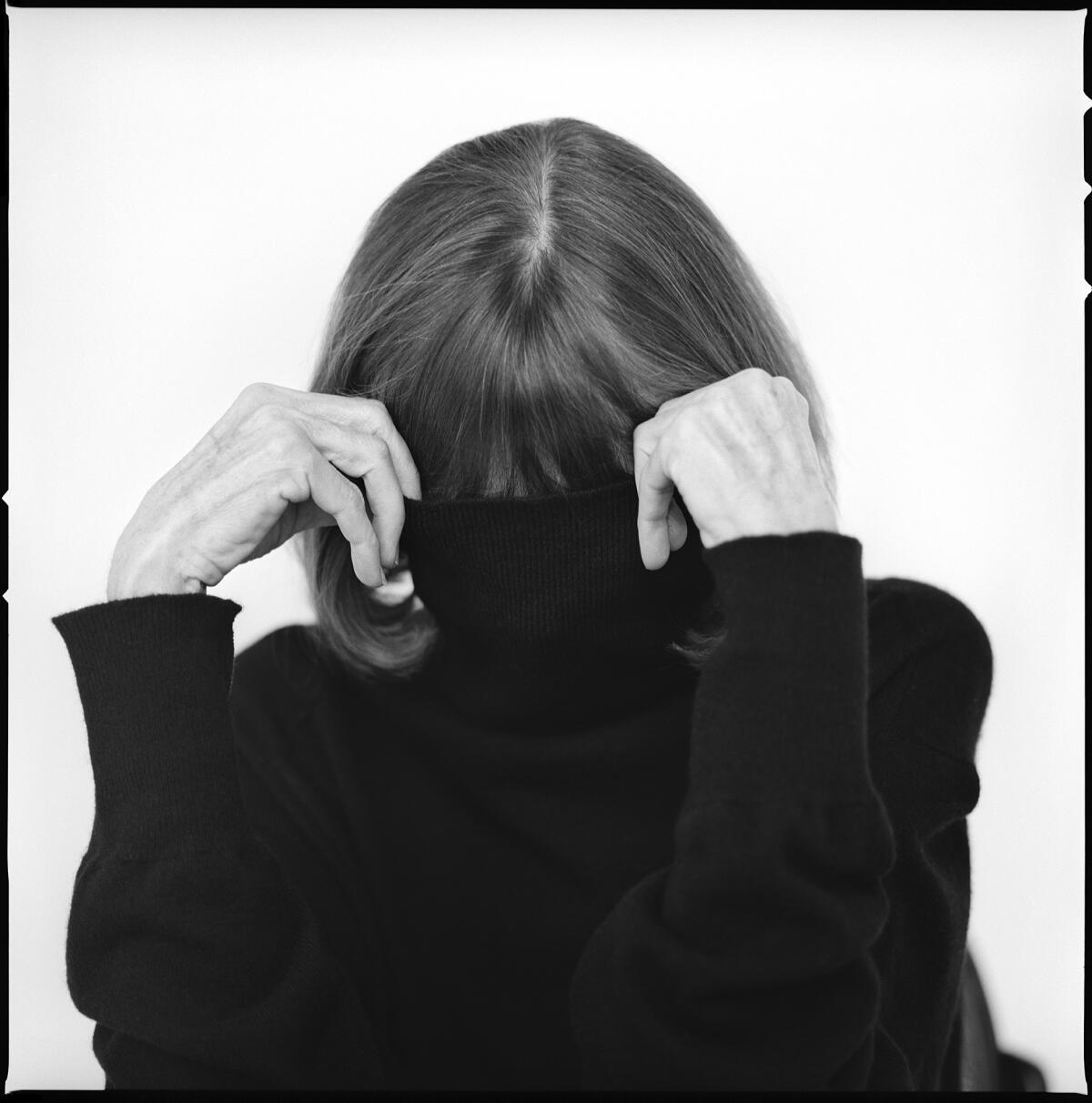 In a black and white image, Joan Didion is scene obscuring her face in her black turtleneck