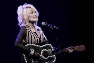 Dolly Parton in a black and beige outfit holding a guitar with pearl detailing and singing on a dark stage