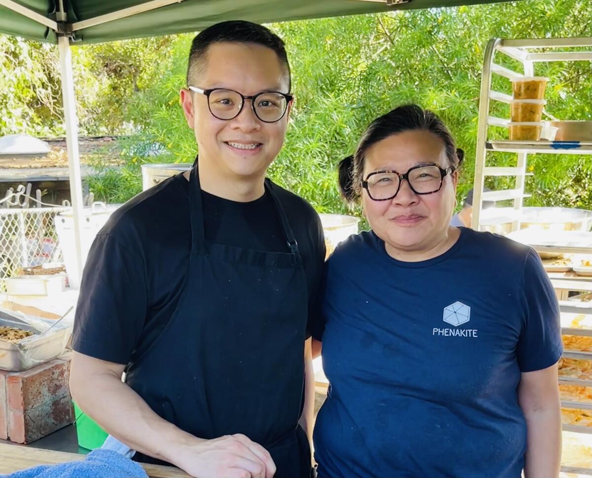 A man and a woman, both wearing T-shirts and dark-rimmed glasses, stand under a tent outdoors.