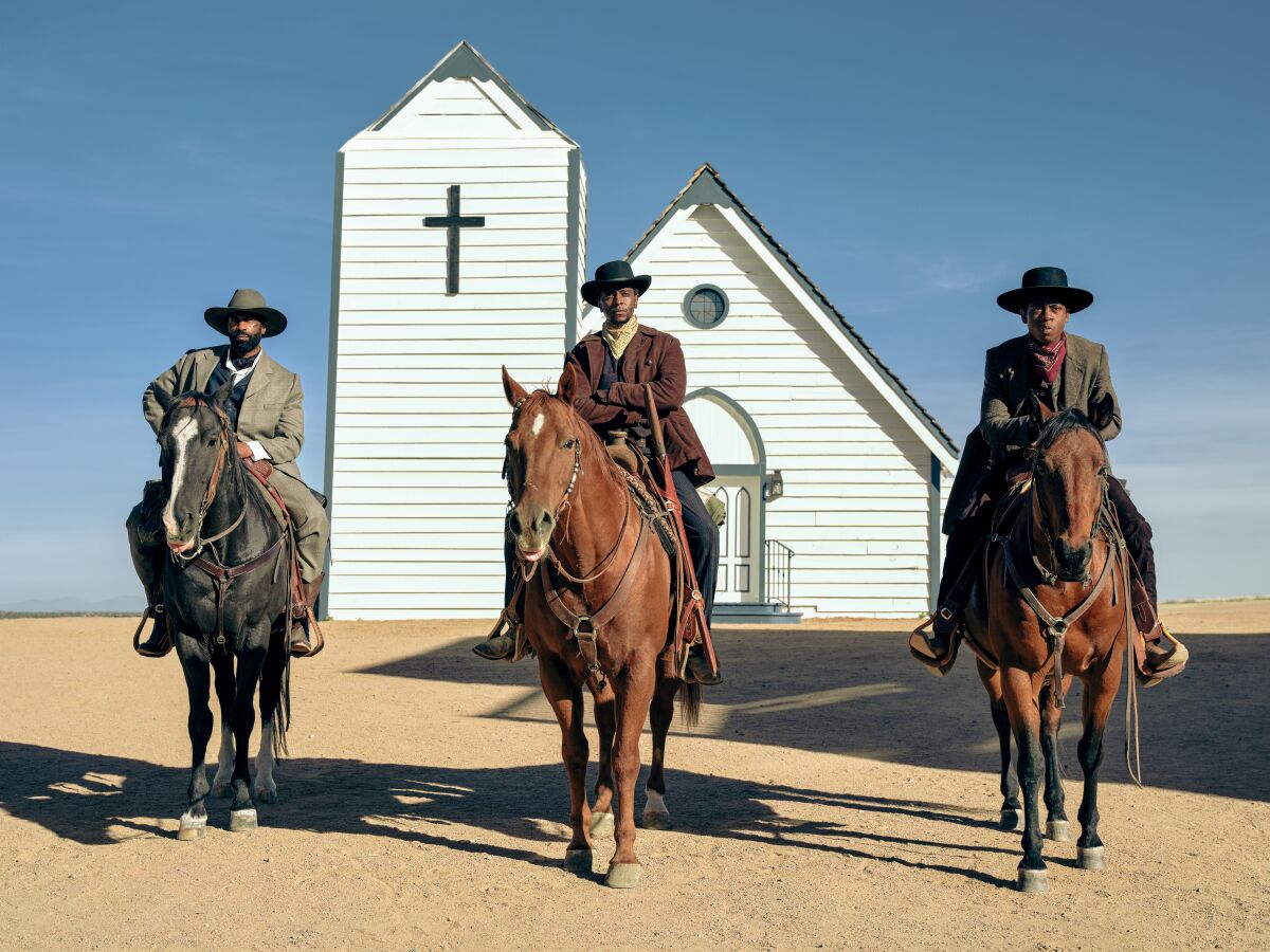 Three men wearing cowboy hats on horses in front of a small white church in the desert.