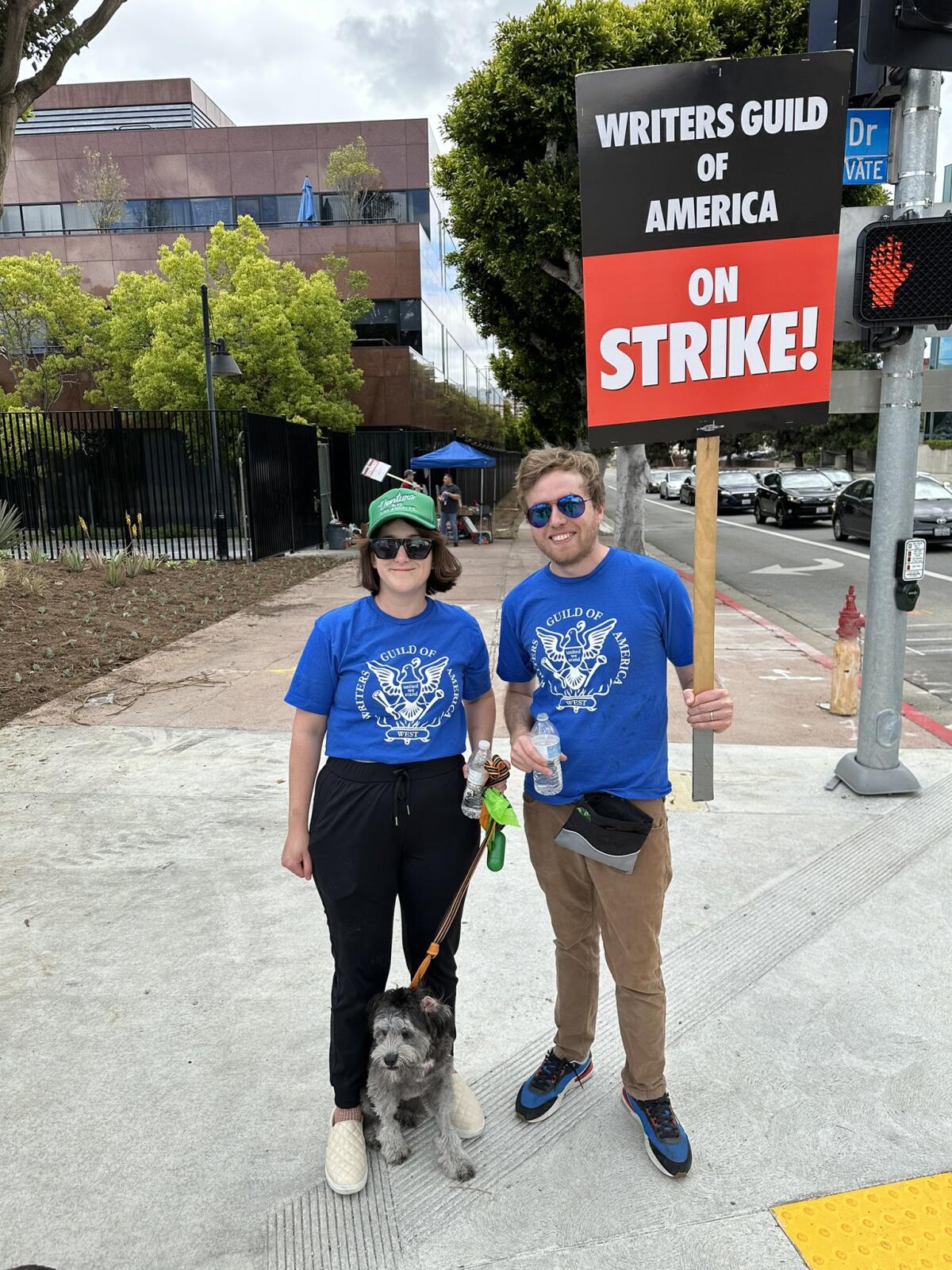 A man and woman stand with their dog on a sidewalk holding a WGA union picket sign