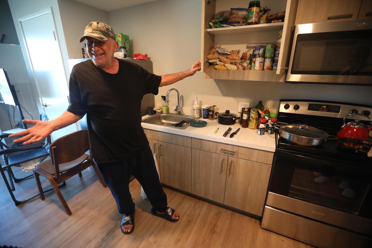 A man in a baseball cap stands next to an open kitchen cabinet.