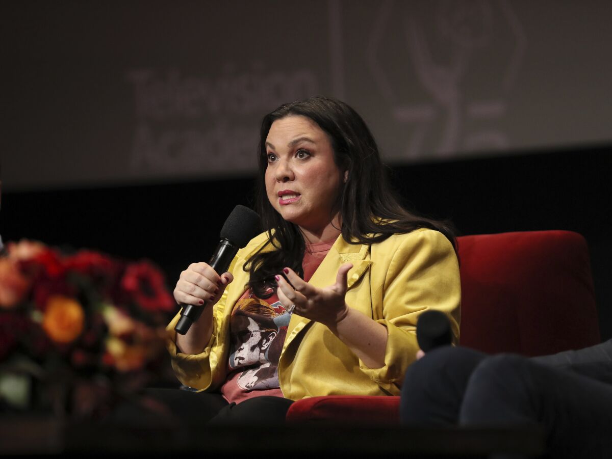 Gloria Calderón Kellett sits on a chair with a microphone in her hand looking toward an audience.
