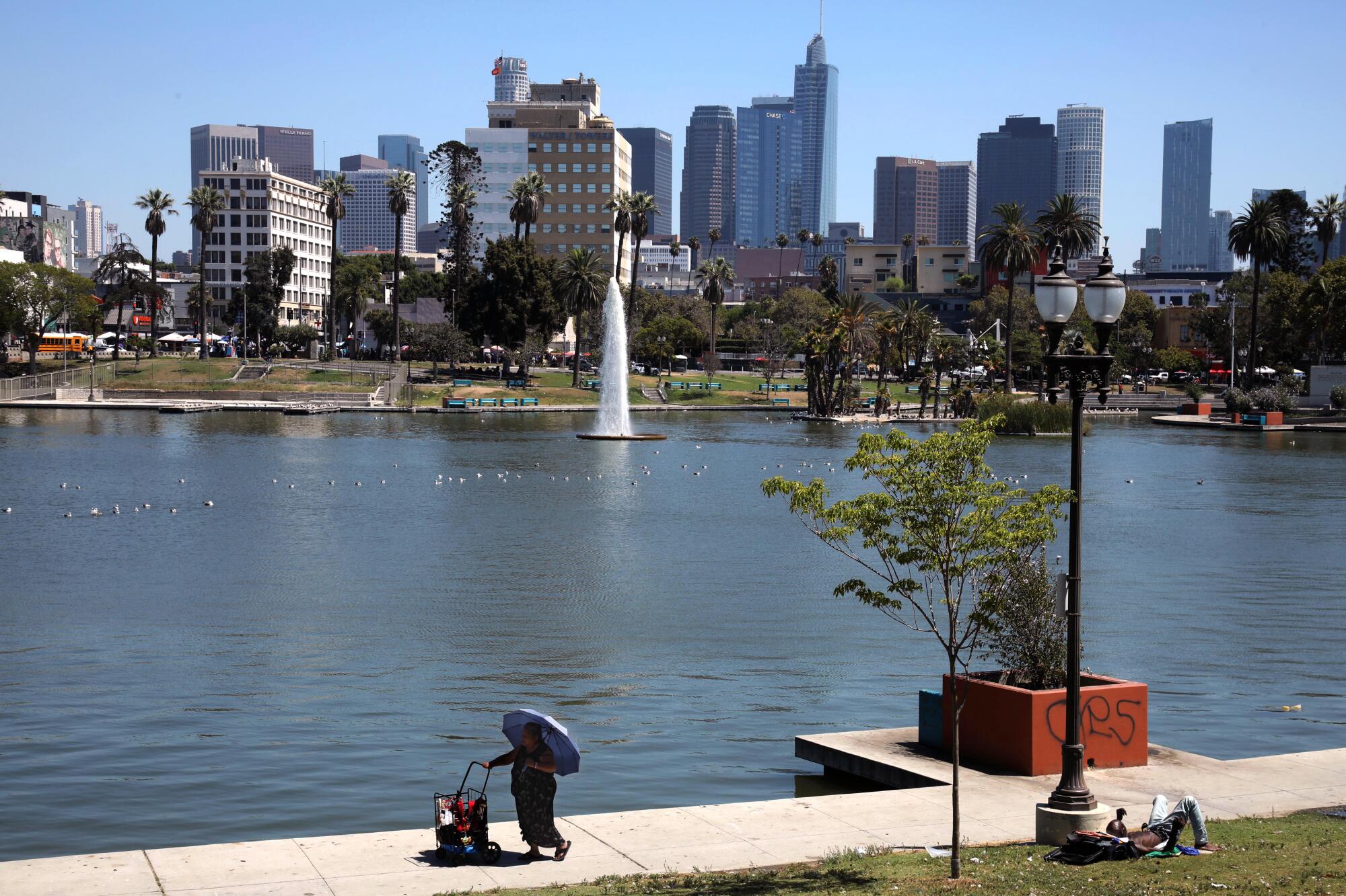 A woman pushes her cart around MacArthur Park Lake against a backdrop of downtown Los Angeles.