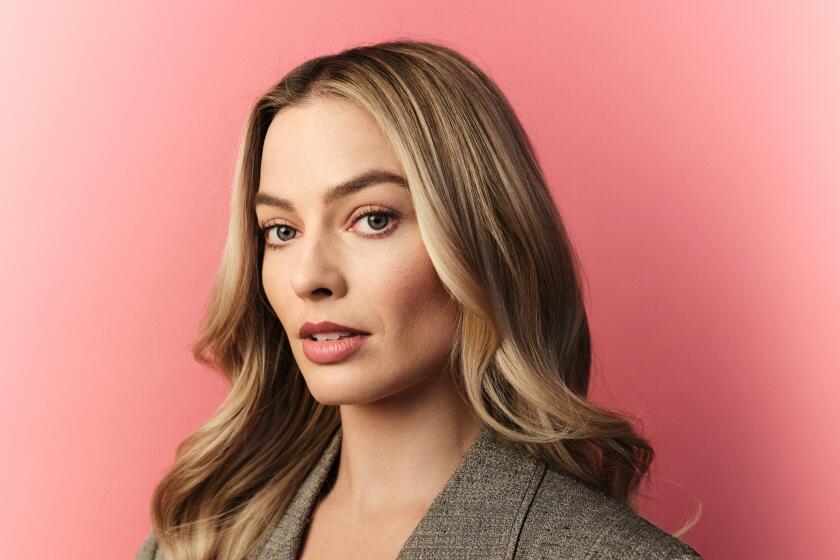 Margot Robbie in a gray oversized blazer, black top standing against a peachy, pink background
